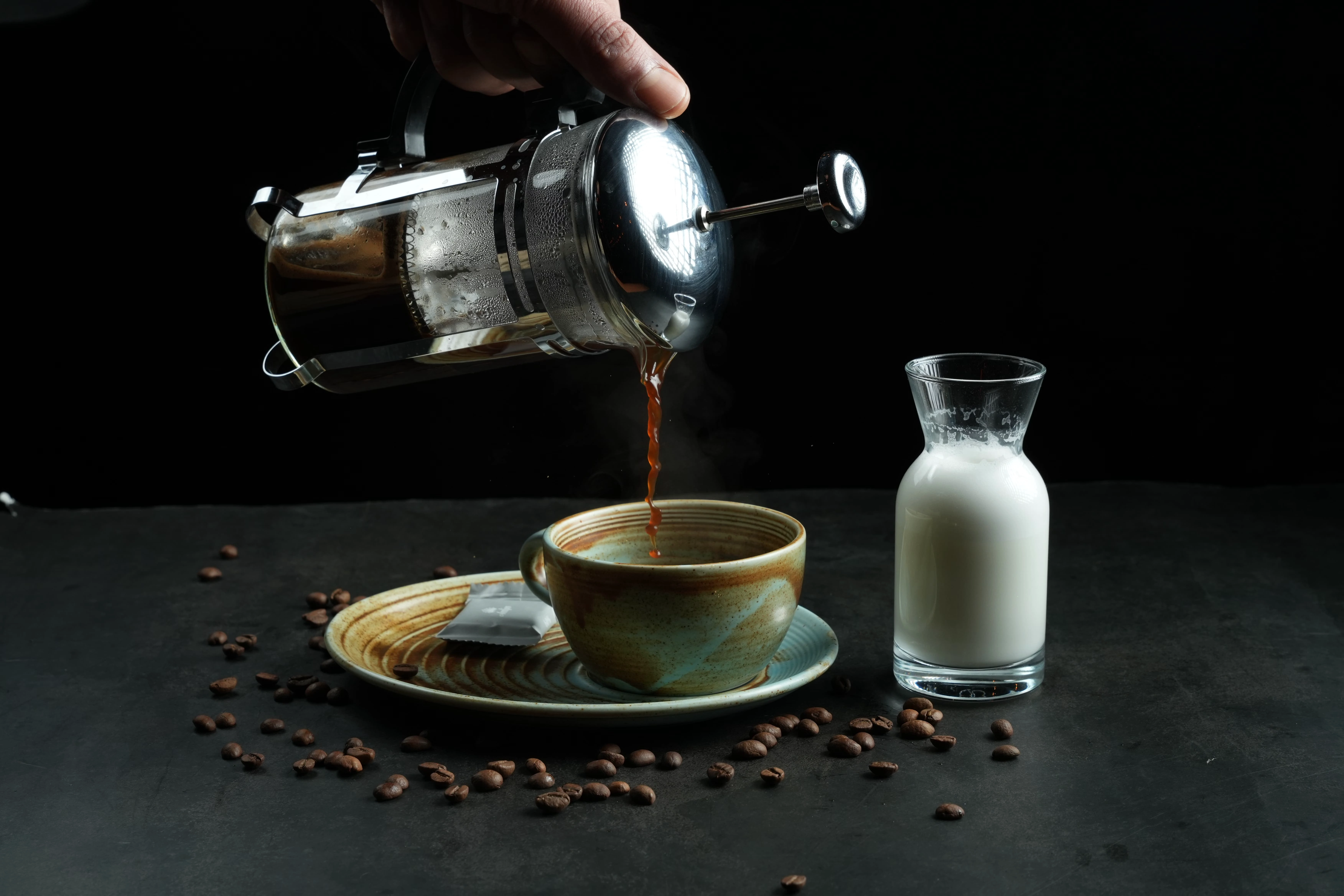Hot French press coffee being poured into a ceramic cup beside a glass bottle of milk
