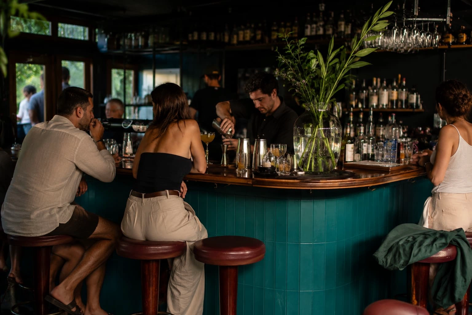Modern teal cocktail bar with guests seated around the counter while a bartender prepares drinks during service