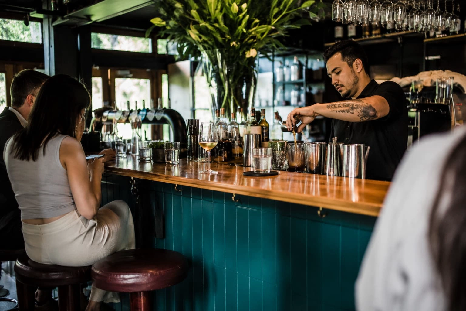 Bartender preparing drinks behind a teal bar counter while guests sit nearby in a modern commercial cocktail bar