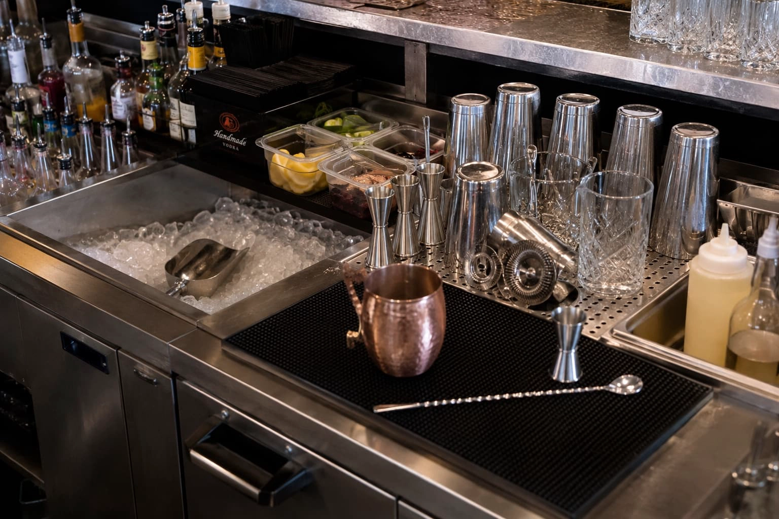 Commercial bar station with an ice well, garnish containers, shaker tins, jiggers, strainers and a separate drink-building surface.