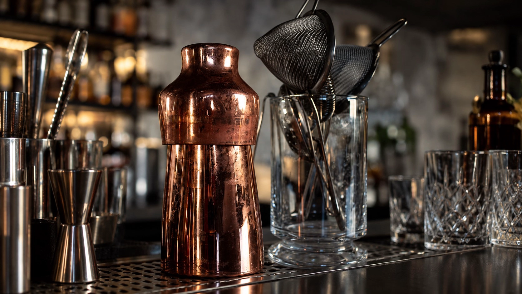 Copper French cocktail shaker displayed on a professional bar counter with strainers, jiggers, and cocktail glasses.